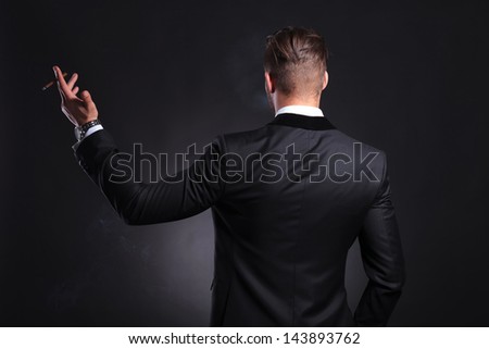 back view of an elegant young fashion man in tuxedo holding a cigar in his raised hand .on black background