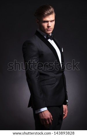 semi profile picture of an elegant young fashion man in tuxedo looking at the camera and frowning. on black background