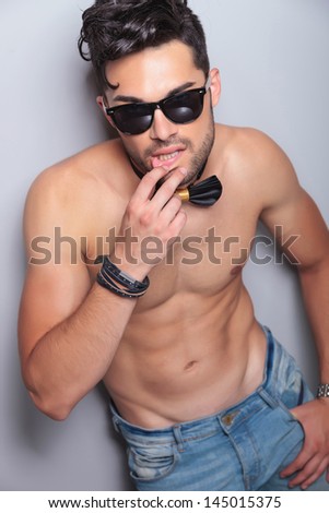 sensual young topless man touching his lower lip and looking at the camera with a thumb in his pocket. on gray background