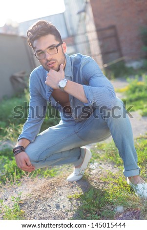 pensive casual young man posing outdoor in a crouched position with his hand at his chin, looking at the camera