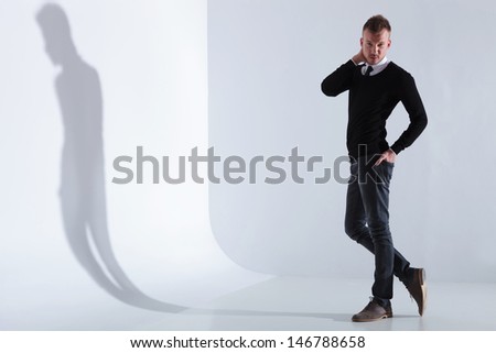 full length picture of a casual young man standing with a hand in his back pocket and the othe r at the back of his head while looking at the camera. on light gray studio background
