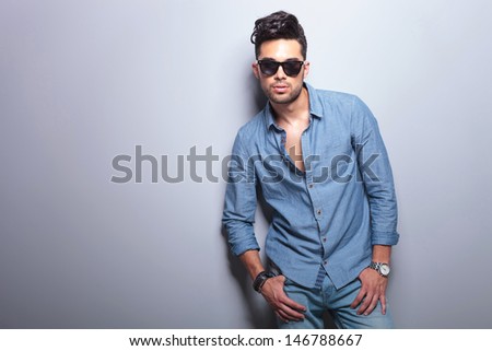 casual young man with both thumbs in pockets poses with his sunglasses on, in the studio. on gray background