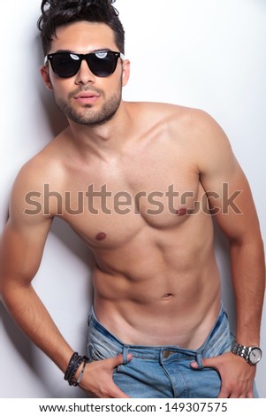 closeup of a young topless man looking at the camera while holding his thumbs in the loops of his jeans. on light gray background
