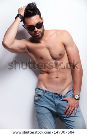 young topless man looking down while passing his hand through his hair and holding his thumb in the loop of his pants. on light gray background