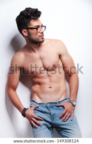 young topless man looking to his side while holding his hands in the loops of his jeans. on light gray background
