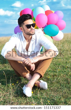 casual young man sitting on the ground with his legs crossed and holding balloons while looking away from the camera