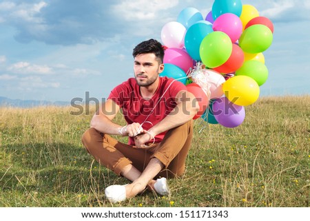 picture of a seated casual young man holding balloons outdoor and looking away from the camera
