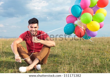 casual young man sitting outdoor on the grass and holding balloons while smiling for the camera