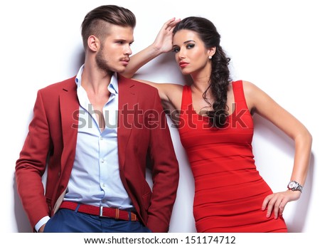 Casual Couple Leaning Against A White Wall, Man With Hands In His Pockets Looking At The Woman . Woman Looking At The Camera While Leaning On His Shoulder