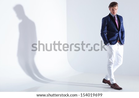 full length portrait of a young business man standing with both hands in his pockets while looking into the camera. on a grau background with shadow