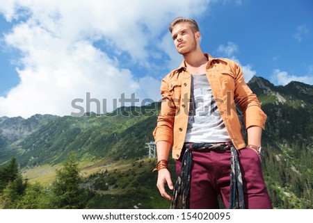 young casual man standing in the mountains, holding a hand in his pocket and looking away from the camera