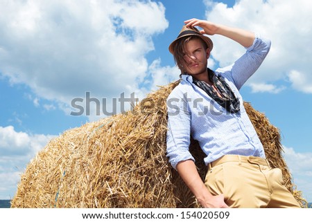 young casual man posing outdoor, leaning on a haystack and looking away from the camera while holding a hand on his hat and a thumb in pocket
