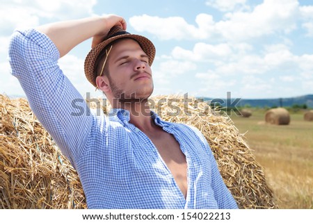 young casual man leaning back on a haystack while holding his hat and a straw behind his ear and his eyes closed