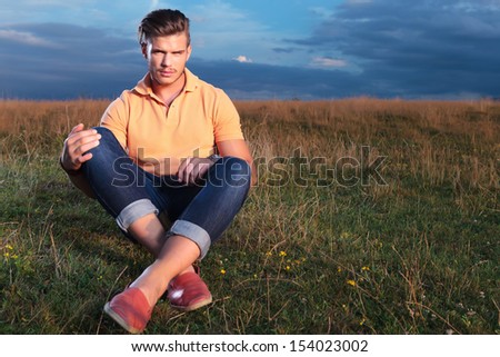 young casual man sitting outdoor on the ground and holding his feet crossed while looking into the camera