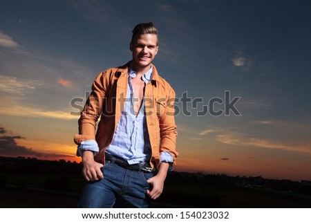 casual young man standing outdoor and holding both thumbs in his pockets while laughing at the camera with the sunset behind
