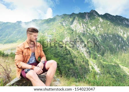 young casual man sitting on a rock, in the mountains and looking away from the camera