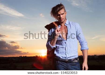 casual young man standing outdoor with his jacket on his shoulder while looking into the camera with the sunset behind him
