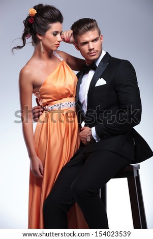 young fashion couple with seated man holding standing woman while looking away from the camera. on gray background