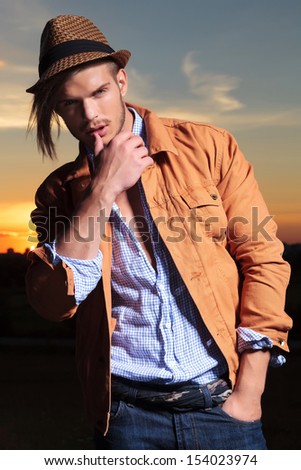 casual young man standing in the sunset with his hand in his pocket and his thumb on his lower lip while looking into the camera