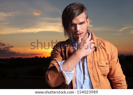 casual young man standing outdoor and touching his lower lip with his thumb while looking into the camera with the sunset behind