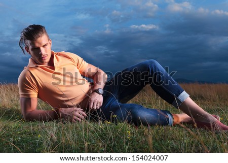 full length photo of a young casual man outdoor laying in the grass and looking into the camera with the dark blue sky behind him