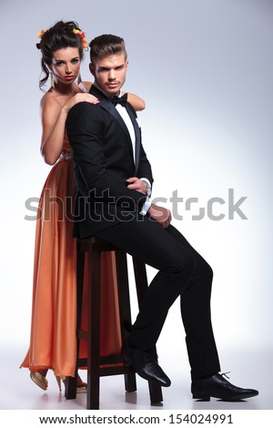 full length photo of a young fashion couple with woman standing with hands on seated man while both looking at the camera. on gray background