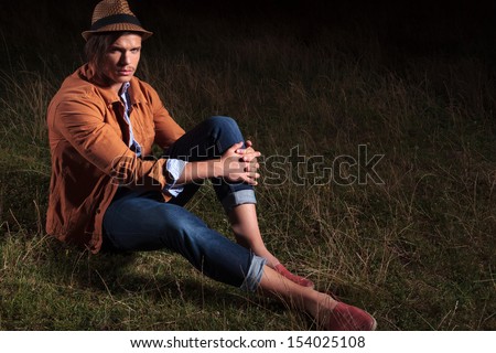 casual young man sitting outdoor and holding his hands on his knee while looking into the camera