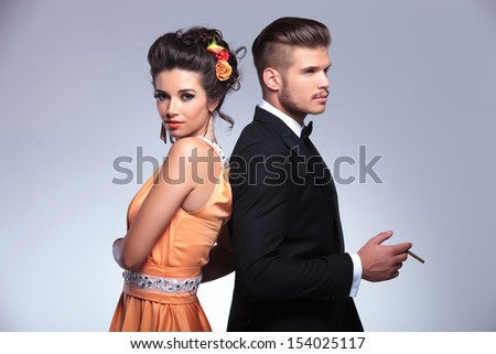 young fashion couple standing back to back while the man is holding a cigarette in his hand. on gray background