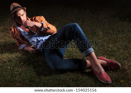full length picture of a casual young man sitting outdoor in the grass and holding a straw in his mouth while looking at the camera