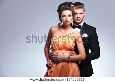 young fashion couple with man behind woman holding her with both hands while both looking at the camera. on gray background