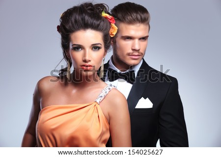 young fashion couple with man behind woman, looking away while she is looking into the camera. on gray background