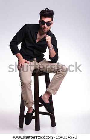full length photo of a young fashion man sitting on a chair and looking into the camera with his eyebrows raised. on a light background