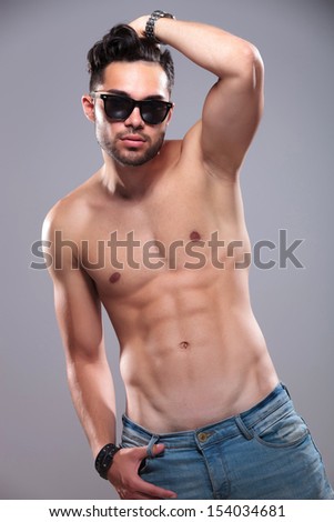 topless young man passing his hand through his hair while holding a thumb in the loop of his jeans and looking into the camera. on gray background