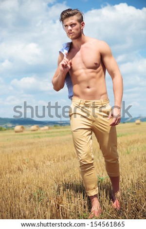 full length portrait of a young topless man walking outdoor with a straw in his mouth while holding his shirt over his shoulder and looking away from the camera