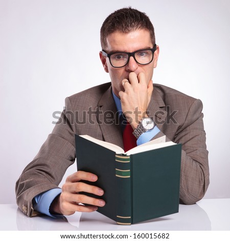 young business man holding a book and reading it frightened, while biting his nails. on a gray background