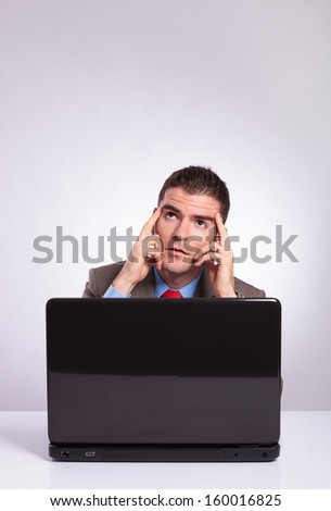 young business man behind his laptop looking upward with his hands on his temples. on a gray background
