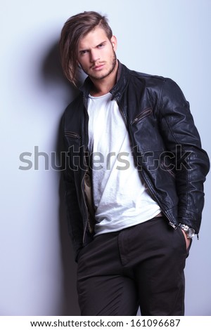 relaxed young man in leather jacket leaning against a gray wall with hands in pockets