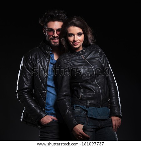 fashion woman in leather jacket standing against her boyfriend, both looking at the camera an smile