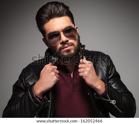 serious bearded fashion man is pulling his jacket\'s collar and looks at the camera