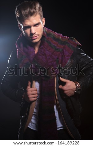 young man adjusting his leather jacket and looking at the camera on dark background