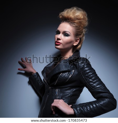 side view of a fashion woman in leather jacket leaning with one hand against a gray studio wall and looking away