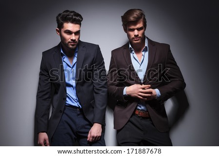two hot male models posing against studio background, one looking away and one unbuttoning his suit