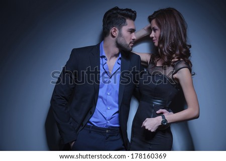 portrait of a young fashion couple looking at each other while the man holds a hand in his pocket and the other on the woman\'s back. on a dark blue background