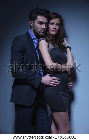 portrait of a young fashion couple where the man holds the woman from behind and looks away over her shoulder and she is looking into the camera. on a dark blue background