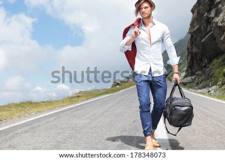sexy young fashion man walking down the middle of a road in the mountains while holding his jacket over his shoulder and a bag in his hand and looking away from the camera