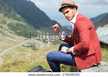 picture of a young fashion man posing outdoor with a cigarette in his hand while sitting on a rock and looking away from the camera, with a beautiful landscape behind him