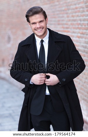 elegant young business man posing outdoor with a big smile on his face while buttoning his suit jacket and looking into the camera