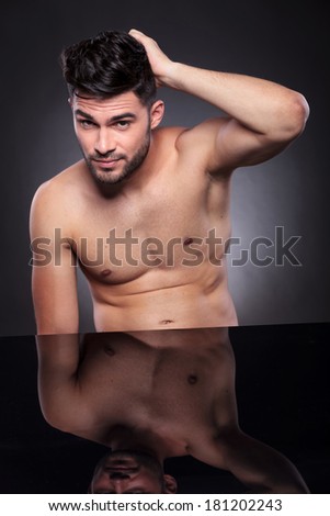portrait of a naked young man sitting at his desk and scratching the back of his head while looking into the camera. on a black studio backgroud
