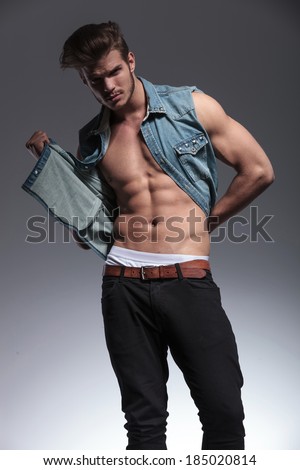 attractive young man pulls his jeans vest and reveals beautiful body while looking into the camera . on a gray background