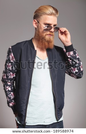 casual young man with a long red beard taking off his sunglasses and looking away from the camera. on gray studio background